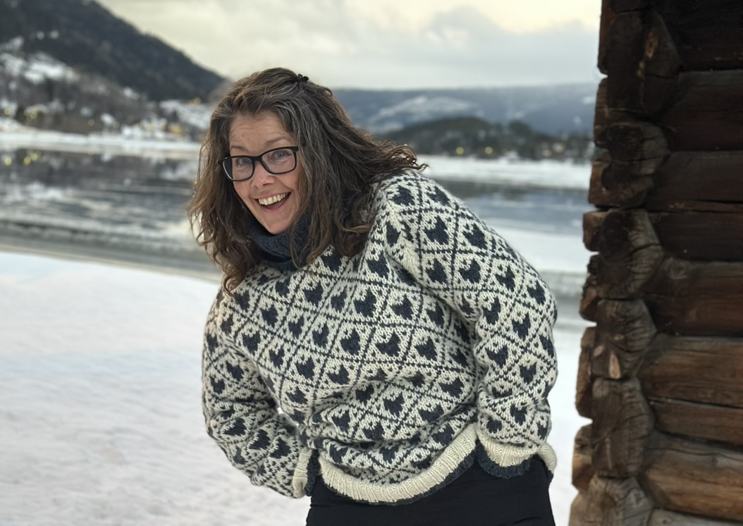 Nærbilde av en smilende kvinne (Marita Aanekre) med briller og langt hår i en tradisjonell norsk strikkegenser med hjertemønster, fotografert utendørs ved en frossen innsjø og en tømmervegg.